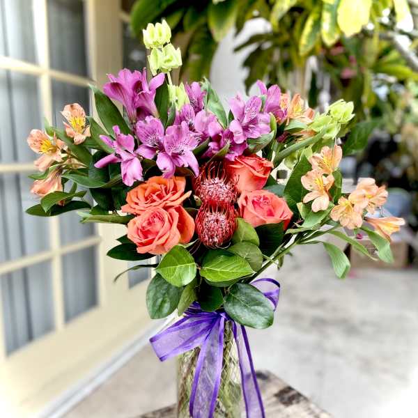 Mixed bouquet of pink and orange flowers in a glass vase with a purple ribbon