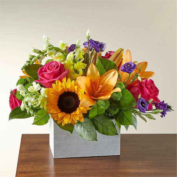 Colorful flower arrangement with roses, sunflower, lilies, and purple blooms in a square vase