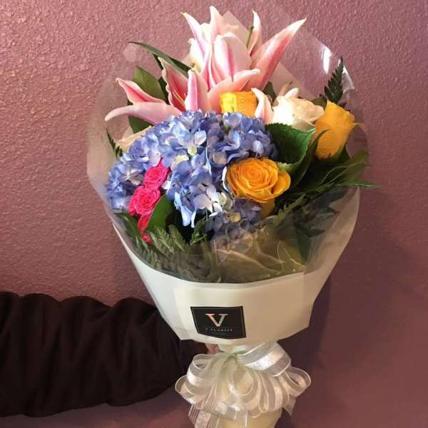 Hand-tied bouquet of pink lilies, blue hydrangea, and yellow and white roses in cream wrap with a bow.