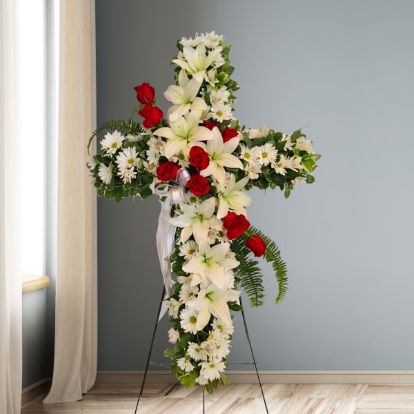 Cross-shaped floral tribute with white lilies, daisies, and red roses on a stand