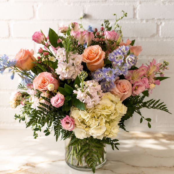 Mixed bouquet of pink, peach, lavender, and white flowers in a glass vase