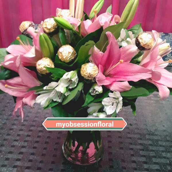 Pink lilies and white flowers arranged in a glass vase with gold-wrapped chocolates