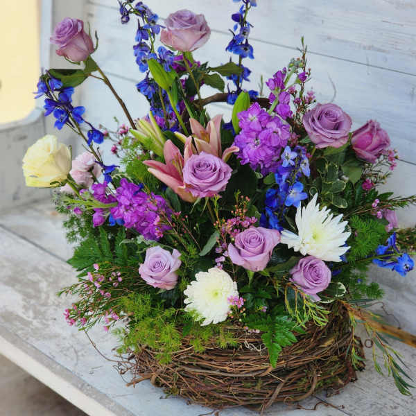 Mixed bouquet of lavender roses, lilies, and blue flowers in a twig basket