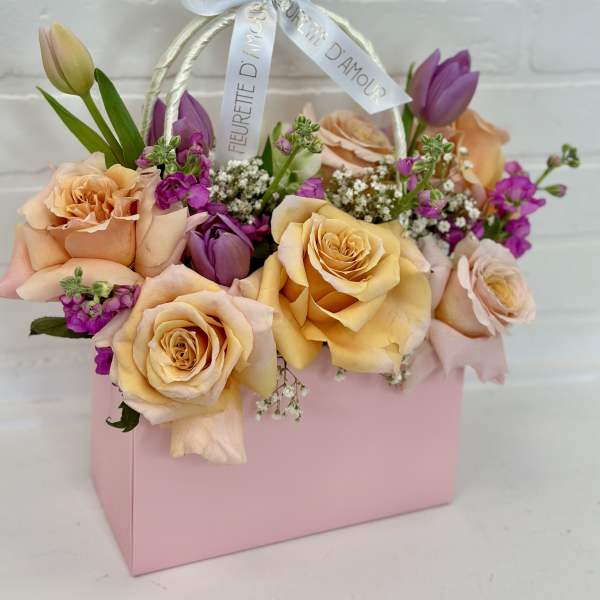 Peach and purple roses arranged in a pink gift box with a ribbon handle.