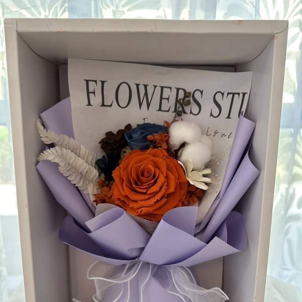 Boxed arrangement with orange rose, blue blooms, and cotton in lavender wrap with white ribbon
