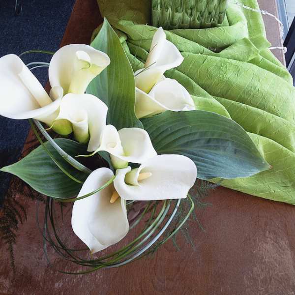 Short arrangement of white calla lilies with large green leaves in a clear bowl on a wooden surface