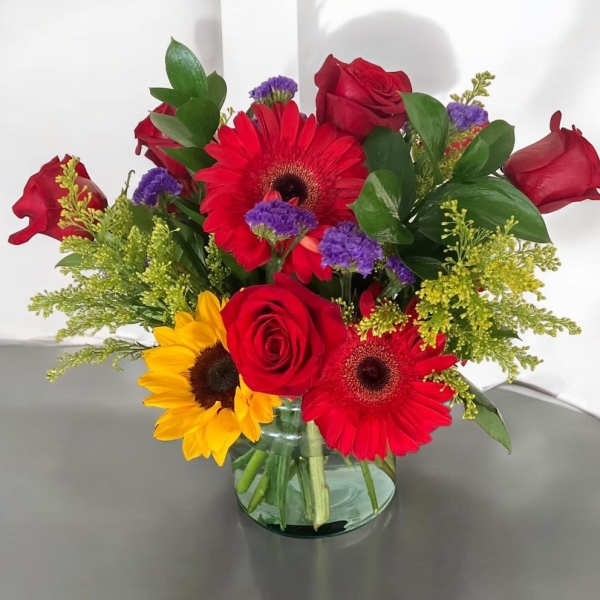 Mixed bouquet of red roses, gerbera daisies, and sunflowers in a glass vase