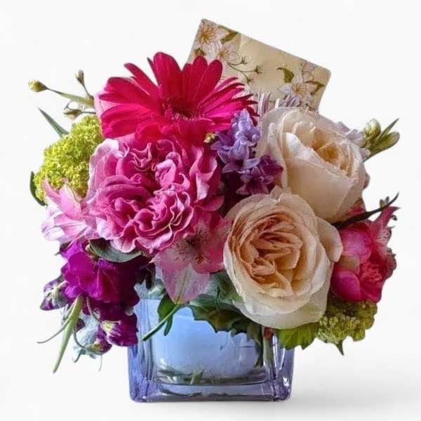 Compact bouquet of pink and cream flowers in a square glass vase with a small card.