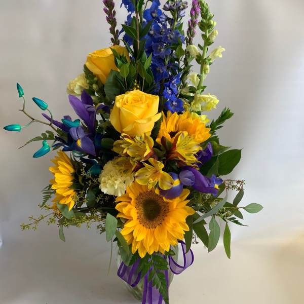 Tall mixed bouquet with sunflowers, yellow roses, and blue flowers in a glass vase