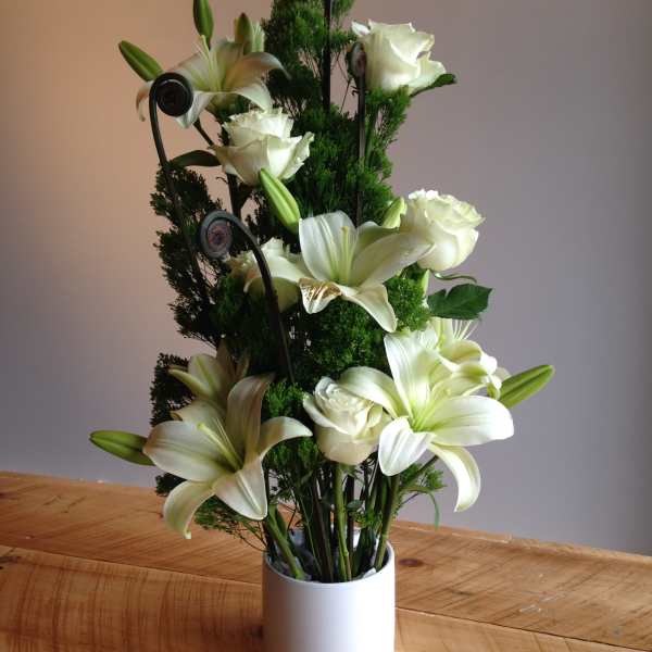 White lilies and roses arranged in a white vase with dark green accents.
