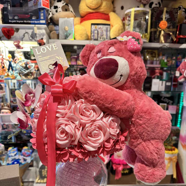 Pink teddy bear hugging a bouquet of pink roses in a glass vase