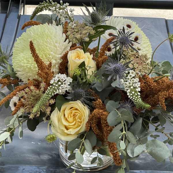 Bouquet of pale yellow roses, white chrysanthemums, and blue thistles in a glass vase