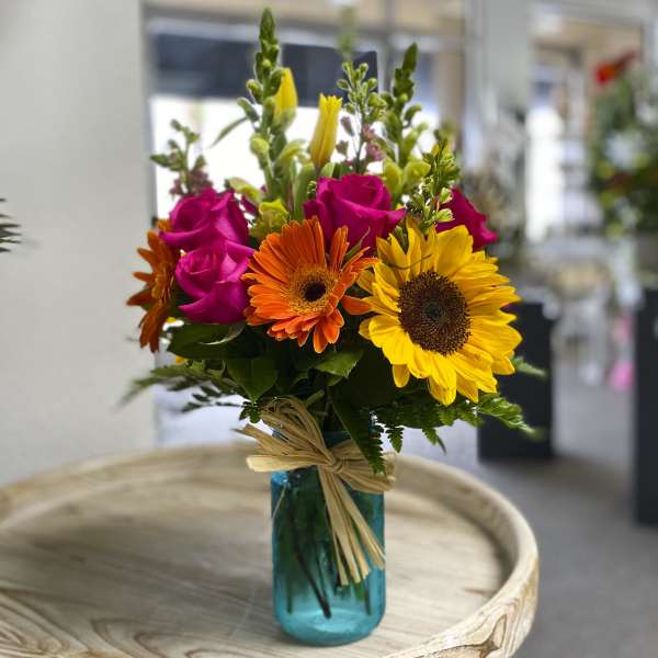 Mixed bouquet of roses, gerbera daisies, and sunflowers in a blue glass vase