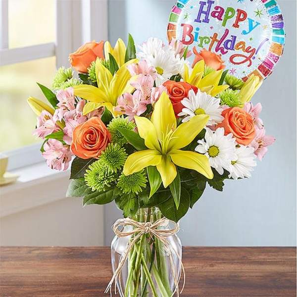 Bouquet of yellow lilies, orange roses, and white daisies in a glass vase with a birthday balloon.