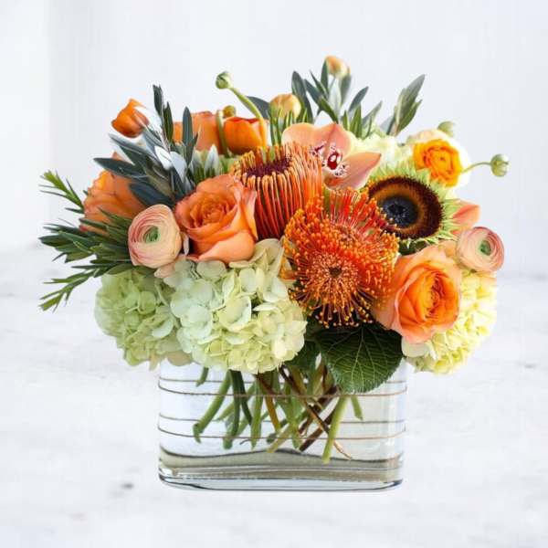 Low orange and green flower arrangement with roses, protea, and hydrangeas in a rectangular clear glass vase