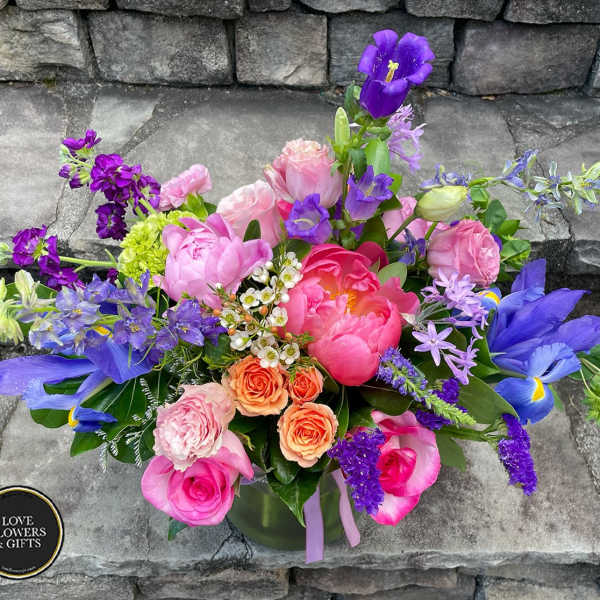 Vibrant mixed arrangement of pink peonies, roses, and purple irises in a clear glass vase