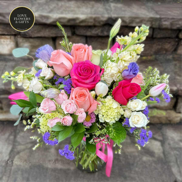 Mixed bouquet of pink, white, and purple flowers arranged in a clear glass vase with a pink ribbon.