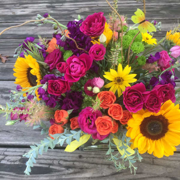 Bright mixed arrangement with sunflowers, hot pink and orange roses in a clear glass vase on a wooden surface