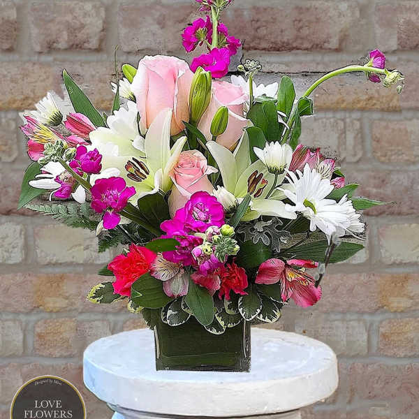 Compact arrangement of pink roses, white lilies and bright pink blooms in a green glass cube vase
