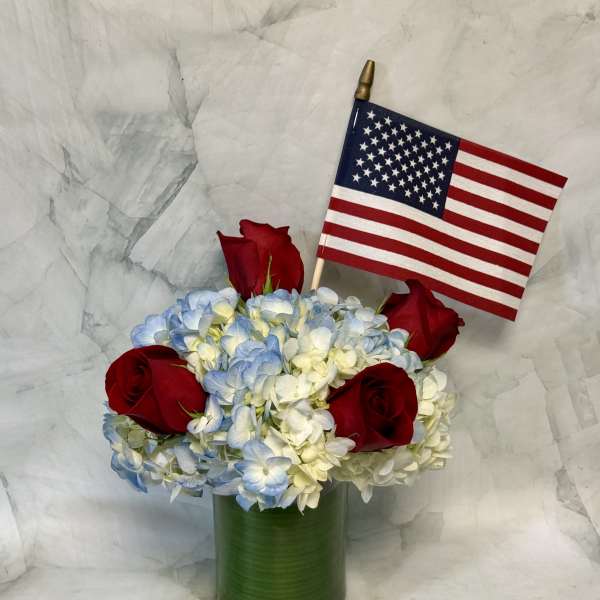 Red roses and blue-white hydrangeas in a green vase with a small U.S. flag