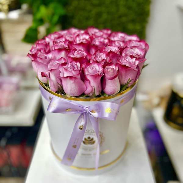 Round white box filled with tightly packed pink roses, finished with a lavender ribbon bow