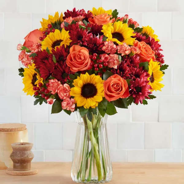 Bouquet of sunflowers, orange roses, and red mums in a clear glass vase