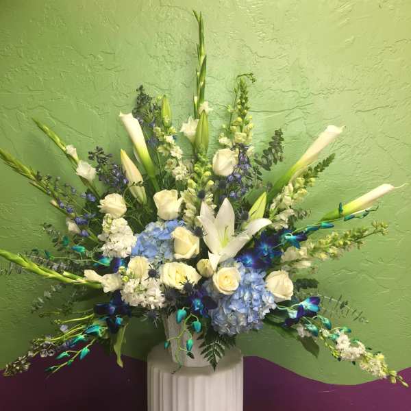 Tall floral arrangement with white lilies, roses, and blue hydrangeas in a white pedestal vase