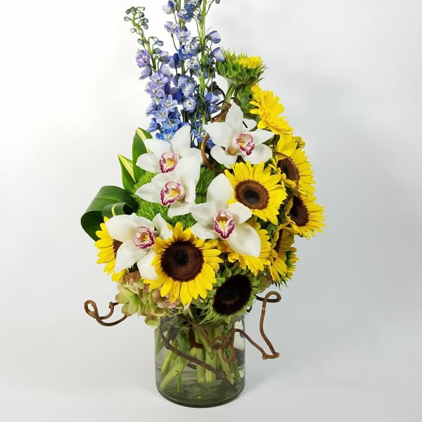 Tall bouquet of sunflowers, white orchids, and blue delphinium in a glass vase