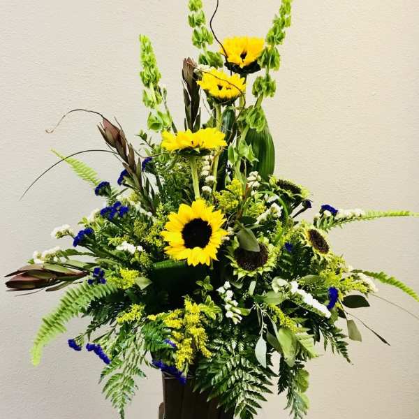 Tall arrangement of yellow sunflowers, green bells of Ireland, and blue accents in a dark rectangular container