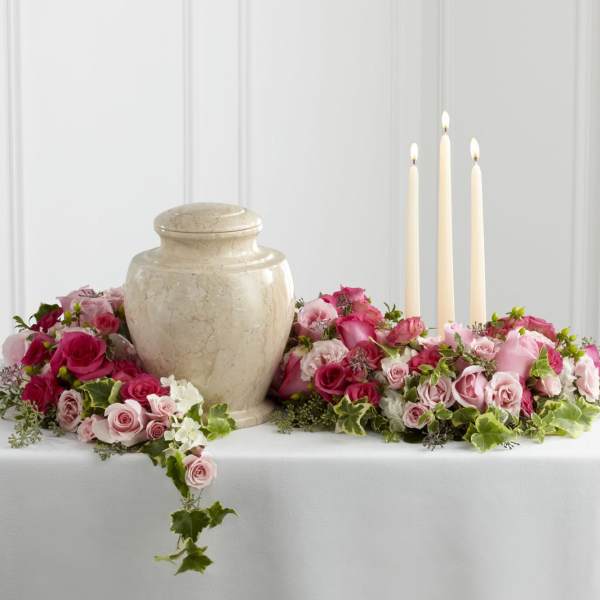 Cremation urn and three ivory candles encircled by pink and white rose flowers on a white table