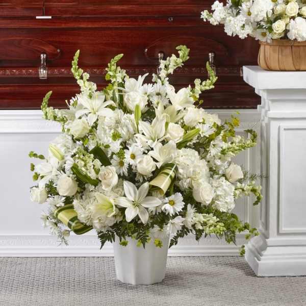 Large white funeral flower arrangement of lilies, roses, and daisies in an urn before a wooden casket