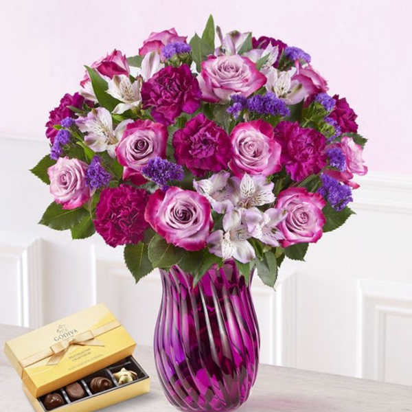 Pink and purple bouquet in a purple glass vase with a box of chocolates