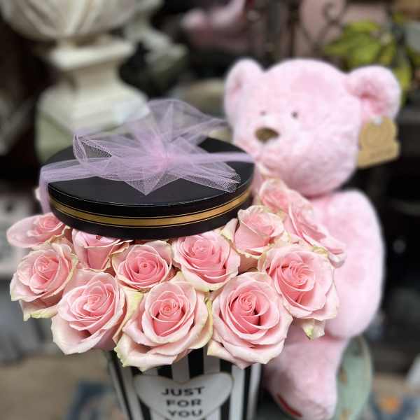 Pink roses arranged in a striped gift box with a teddy bear behind it