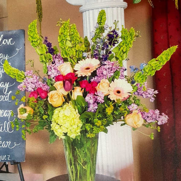 Tall mixed arrangement of pink, peach, and green flowers in a clear glass vase on a table