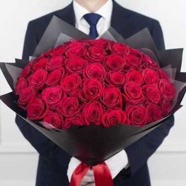 Large bouquet of tightly packed red roses wrapped in black paper, held by a person in a dark suit.