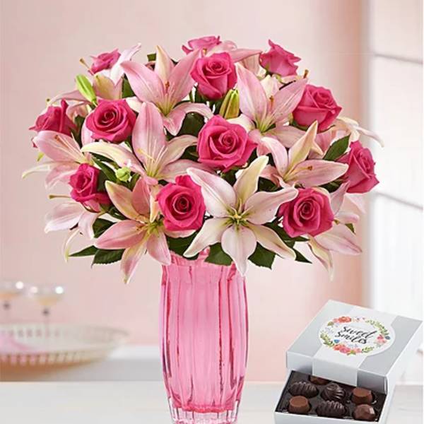 Pink roses and lilies in a pink glass vase beside a box of chocolates