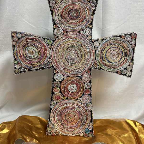 Multicolor decorative cross made of coiled circular patterns, set against white fabric with gold cloth at the base.