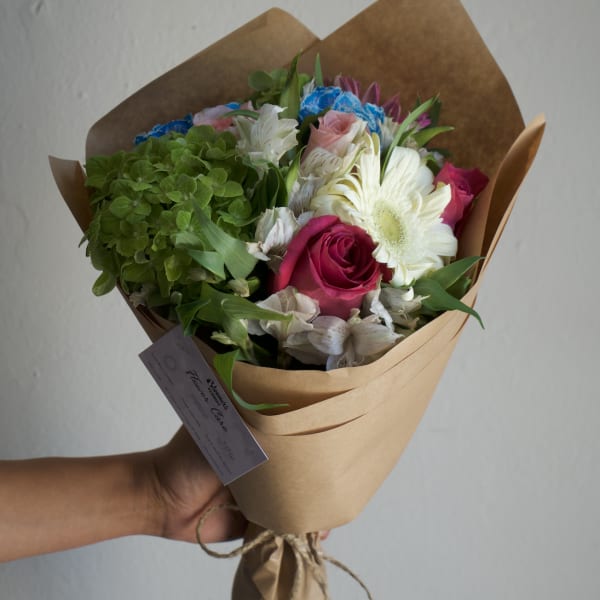 Handheld bouquet with pink roses, white gerbera daisies, and green hydrangea in brown paper