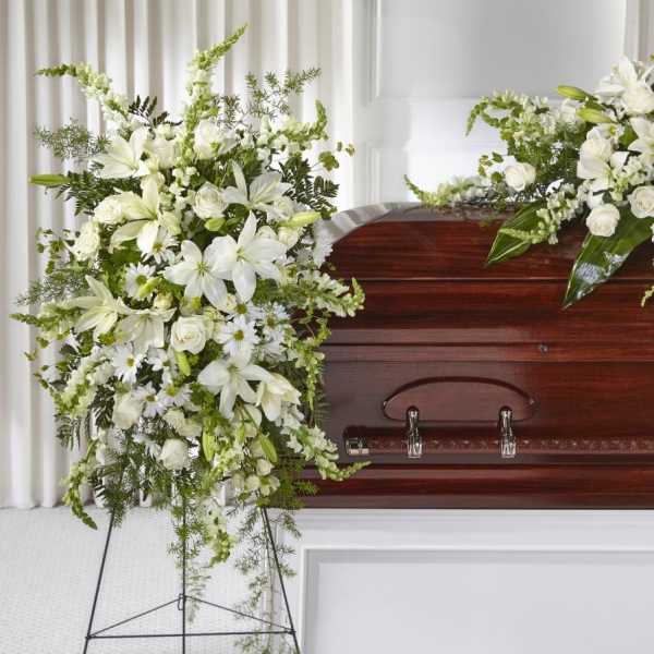 Standing spray of white lilies, roses, and daisies beside a wood casket in a funeral setting