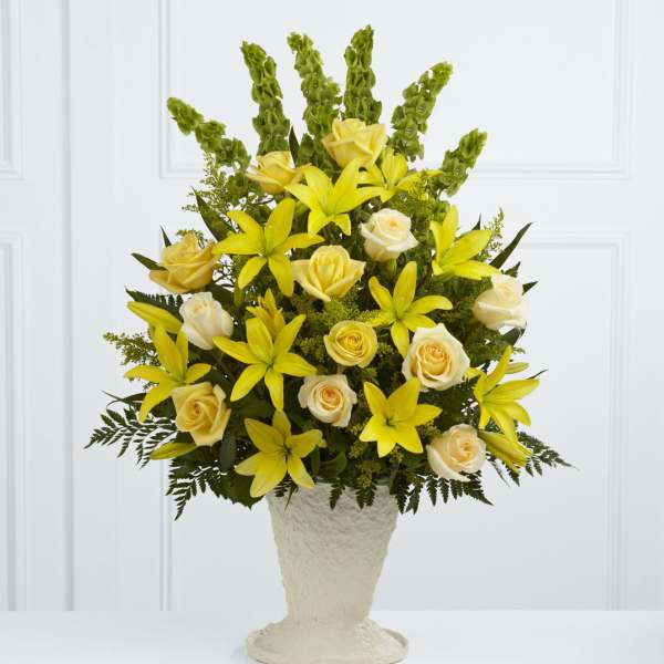 Tall yellow lilies and cream roses in an ivory pedestal vase with vertical green accents