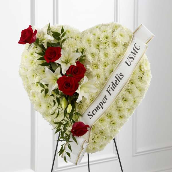 Heart-shaped white mum funeral spray with red roses, white lilies, and a tribute ribbon on an easel