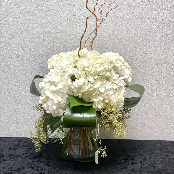 White hydrangea arrangement with structured green leaves in a clear glass vase and curly willow branches