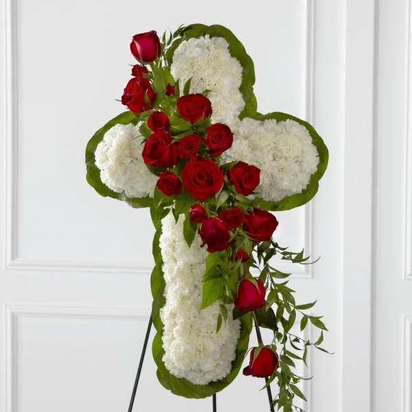 Standing cross of white flowers with red roses and cascading greenery on a metal easel.