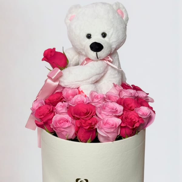 White teddy bear sitting in a box of pink roses with a ribbon
