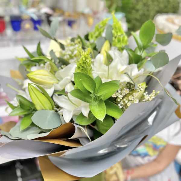 Hand-tied bouquet of white lilies and small white flowers with vivid green accents wrapped in gray and gold paper.