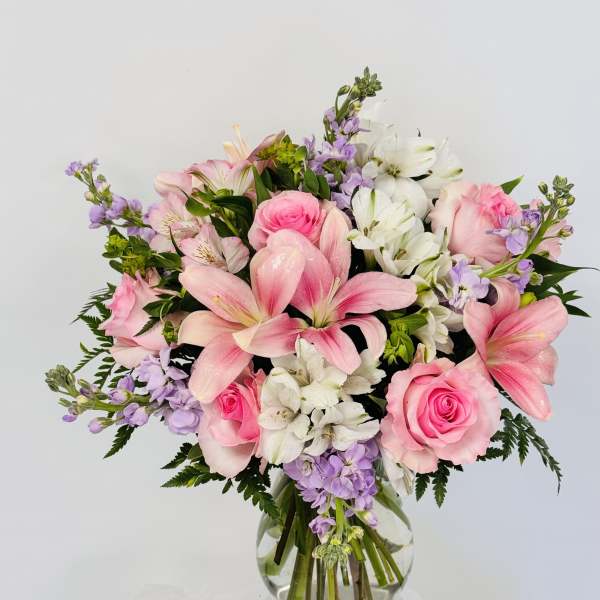 Pink lilies and roses arranged in a clear glass vase with white and lavender flowers