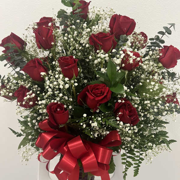 Red roses arranged in a vase with white baby's breath and a red ribbon bow.