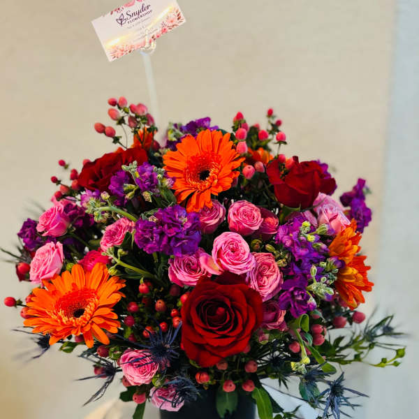 Bright mixed bouquet with red roses, pink spray roses, orange gerberas, and purple blooms in a black vase