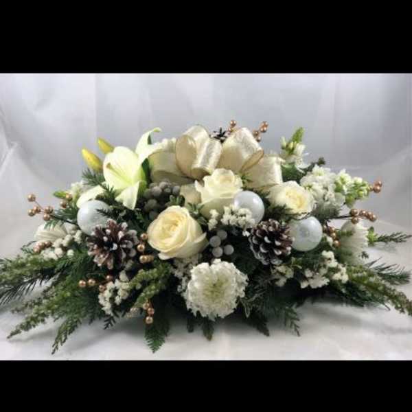 Low white flower centerpiece with pinecones, ornaments, and a gold bow in winter evergreens