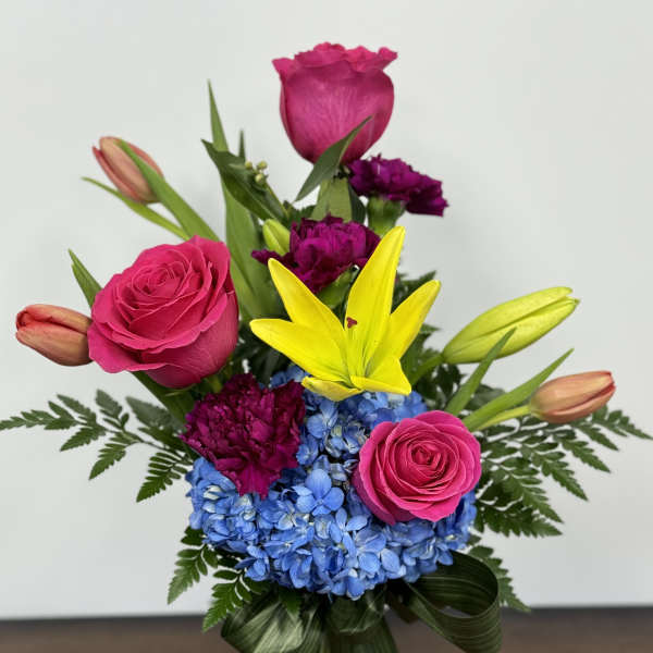 Bright bouquet with pink roses, yellow lilies, blue hydrangea, and purple carnations in a glass vase